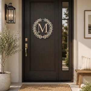 Può includere: Una porta d'ingresso in legno marrone scuro con una decorazione a ghirlanda circolare in legno chiaro al centro. La ghirlanda circonda una grande lettera M maiuscola in carattere serif. La porta include un pannello di vetro verticale e maniglie dorate.