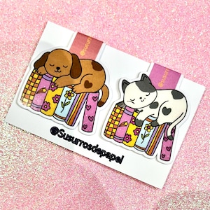 Puede incluir: Dos marcapáginas magnéticos con un perro marrón y un gato blanco y negro durmiendo sobre una pila de libros ilustrados coloridos. Los marcapáginas están sujetos a una tarjeta blanca con el texto @Susurrosdepapel impreso en negro.
