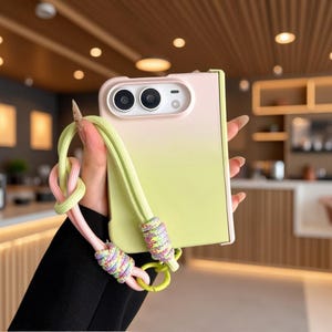 May include: A foldable smartphone case featuring a soft gradient transition from pale pink at the top to light lime green at the bottom. The case includes a matching lime green and pink knotted rope lanyard attached with decorative multi-colored cord wraps.
