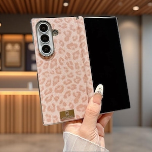 以下が含まれることがあります： ライトピンクとベージュのヒョウ柄が施された長方形のスマートフォンケース。ケースには3つの円形レンズを備えた縦型のカメラ用切り抜きがあり、下部中央には小さな長方形のゴールドトーンの金属プレートが付いています。