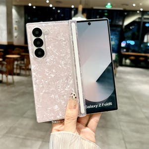 May include: A light pink, pearlescent, textured phone case for a Galaxy Z Fold6. The case features a glossy, marbled finish with a dedicated slot for a stylus pen. The phone screen is visible, displaying the text Galaxy Z Fold6 at the bottom.