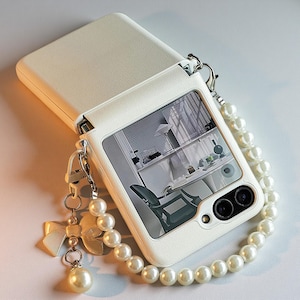 May include: A white foldable smartphone case with a textured finish, featuring a pearl bead wrist strap and a dangling charm shaped like a bow with a single pearl. The case displays a black and white interior room scene on its outer screen.