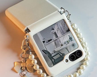 May include: A white foldable smartphone case with a textured finish, featuring a pearl bead wrist strap and a dangling charm shaped like a bow with a single pearl. The case displays a black and white interior room scene on its outer screen.