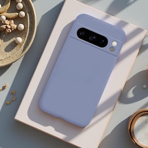 May include: A light periwinkle blue smartphone case with a smooth, matte finish. The case features a rectangular camera cutout at the top, exposing a black camera module with multiple lenses and a circular flash component on the right side.
