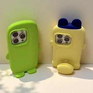 Op de afbeelding: Twee siliconen telefoonhoesjes met camera-uitsparingen. Het linkerhoesje is effen limoengroen met kleine voetjes. Het rechterhoesje is lichtgeel met donkerblauwe oren aan de bovenkant en een ronde standaard aan de onderkant.