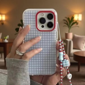 May include: A clear smartphone case with a light blue and white gingham pattern, featuring a red camera lens border. Attached is a beaded phone charm with red, light blue, and white beads, including a square gingham bead and a white heart charm.