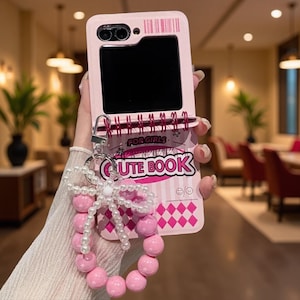 以下が含まれることがあります： スパイラル綴じのノートを模したライトピンクの折りたたみ式スマホケース。ケースには「FOR GIRLS」と「CUTE BOOK」の文字がグラフィック調で描かれており、ピンクのビーズと白いパールのリボンで作られた装飾的なリストストラップが付いています。