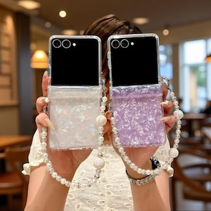 May include: Two rectangular phone cases with black upper sections and lower sections featuring iridescent, crushed-look textures in white and light purple. Each case includes a long, decorative strap made of white faux pearls and silver beads.