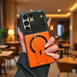 Puede incluir: Una funda rectangular para smartphone con una sección negra para el módulo de cámara y una sección inferior de color naranja vibrante. La parte naranja incluye un anillo magnético circular y un soporte plegable centrado en la parte trasera.