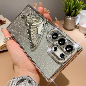 Könnte beinhalten: Eine rechteckige, silber glitzernde Handyhülle mit einem großen, dreidimensionalen, metallisch silbernen Schmetterlingsornament auf der Rückseite. Der Kameraausschnitt ist mit kleinen, klaren Kristallen verziert und die Kanten haben ein silbernes Finish.