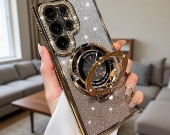 Puede incluir: Una funda rectangular para smartphone con acabado de purpurina plateada y bordes metálicos dorados. La funda cuenta con un soporte de anillo circular dorado en el centro con el texto KATHY TECH y un recorte para la cámara con borde dorado.
