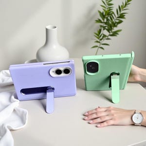May include: A light purple foldable smartphone case and a light green tablet case, both featuring integrated kickstands. The purple case has a horizontal camera cutout, while the green case has a square camera cutout with rounded corners.