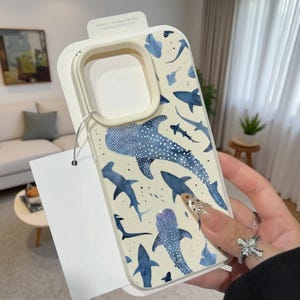 May include: A protective phone case featuring a cream-colored background with a repeating pattern of blue whale sharks and various shark silhouettes. The case includes a raised cream-colored camera frame and a white paper tag attached by a silver string.
