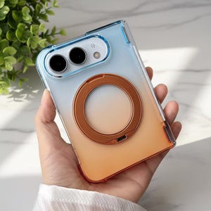May include: A clear smartphone case with a blue-to-orange gradient finish, featuring a prominent circular metallic copper-colored magnetic ring attachment on the back and a cutout section for the camera lenses and flash.