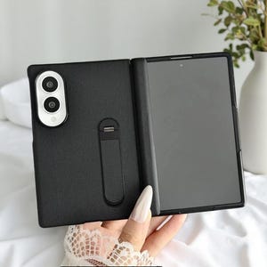 Könnte beinhalten: Eine schwarze, faltbare Smartphone-Hülle mit strukturierter Oberfläche, die einen vertikalen Kameraausschnitt mit zwei Linsen und silberner Umrandung sowie einen rechteckigen integrierten Ständer auf der Rückseite und ein dunkles Display besitzt.