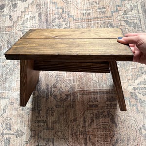 May include: A small, rectangular wooden stool with a dark brown stained finish. The stool features a flat top surface constructed from two joined planks and two angled side supports that provide a stable base for this functional furniture piece.