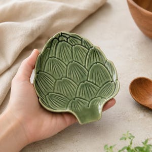 Puede incluir: Un pequeño plato de cerámica verde con forma de alcachofa. La superficie presenta un patrón texturizado en relieve de hojas superpuestas con un acabado brillante. El plato tiene una forma redondeada y orgánica que imita a una alcachofa fresca.