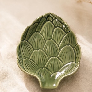 May include: A ceramic dish shaped like an artichoke, featuring a glossy, sage green glaze with embossed, overlapping leaf patterns that create a textured, scalloped edge. The design mimics the natural layers of an artichoke bulb in a compact form.