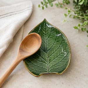 May include: A glossy green ceramic spoon rest shaped like a leaf with a metallic gold rim, featuring embossed vein details on the surface. A light brown wooden spoon with a smooth, rounded bowl rests diagonally across the center of the leaf dish.