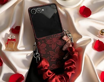 May include: A foldable smartphone case featuring a dark red floral lace pattern on the lower half and a black upper section with two camera lenses. The case includes a detachable, gathered red satin wrist strap attached with silver metal clips.