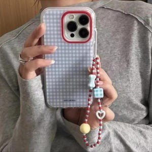 May include: A light blue and white gingham patterned phone case with a red camera frame border. Attached is a beaded phone strap featuring small red beads, a light blue bone-shaped bead, a blue gingham cube bead, a white heart bead, and a yellow sphere.