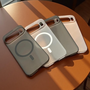 Può includere: Quattro cover per smartphone disposte in fila, caratterizzate da un design con anello di ricarica magnetico sul retro. Le cover sono disponibili in grigio scuro traslucido, bianco semitrasparente, grigio scuro solido e beige chiaro o bianco sporco.