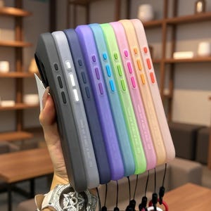 May include: A collection of eight silicone phone cases arranged in a row, featuring a spectrum of colors including black, light gray, dark blue, purple, teal, lime green, pink, and peach. Each case has matching side buttons and a small black wrist strap.