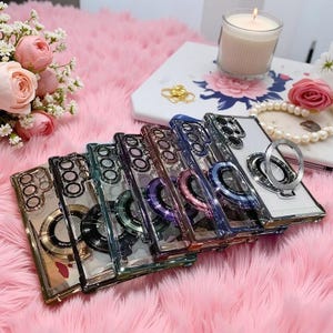May include: A collection of six transparent smartphone cases arranged in a row, each featuring a metallic camera lens frame and a circular ring stand on the back. The cases come in various metallic colours including gold, black, green, purple, pink, and blue.