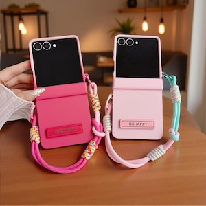 Puede incluir: Dos fundas plegables para smartphone con acabado mate, una en rosa oscuro y otra en rosa claro. Cada funda presenta una placa rectangular con el texto QGHAPPY y una larga correa de cuerda trenzada multicolor unida a los enganches laterales.