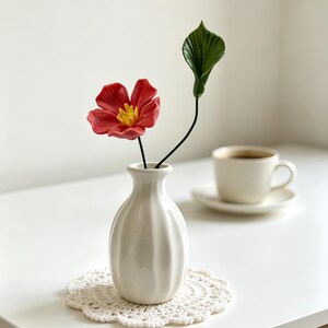 May include: A small white ceramic vase with vertical fluting holds a single red flower with a yellow center and a separate green leaf on a thin stem. The vase sits on a round white crocheted doily, showcasing a clean and simple aesthetic.