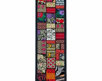 Pode incluir: Um caminho de mesa retangular com um design de retalhos de vários quadrados de tecido bordados em cores vibrantes, incluindo vermelho, amarelo, verde, roxo e preto. O caminho é contornado com tecido preto e finalizado com borlas pretas.