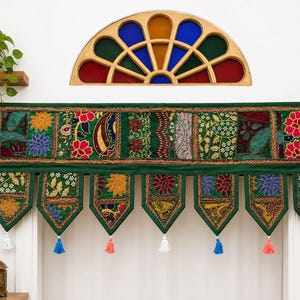May include: A rectangular green fabric valance featuring a patchwork of embroidered floral and paisley patterns in red, yellow, blue, and white. The valance has seven pointed hanging tabs, each finished with a small white, blue, or pink tassel.