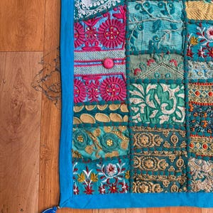 Pode incluir: Uma tape&ccedil;aria retangular com um mosaico de quadrados de tecido bordados em tons de turquesa, azul-petr&oacute;leo, magenta e dourado. A pe&ccedil;a &eacute; contornada por um acabamento de tecido azul s&oacute;lido e inclui uma pequena borla azul no canto inferior.