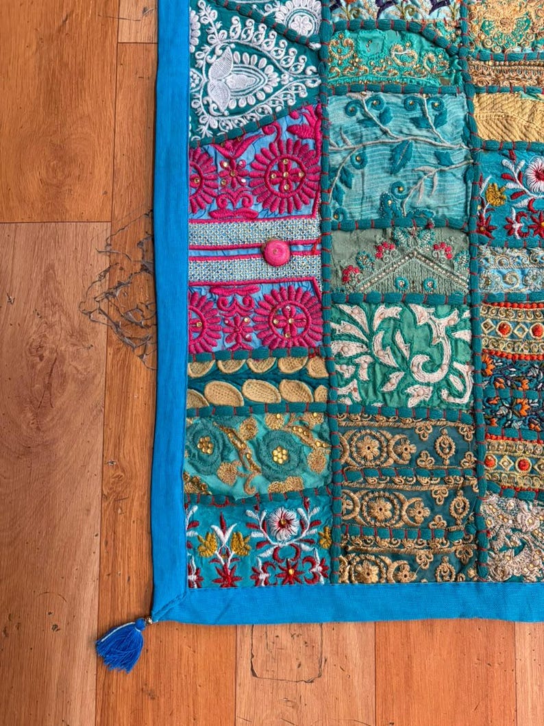 Pode incluir: Uma tape&ccedil;aria retangular com um mosaico de quadrados de tecido bordados em tons de turquesa, azul-petr&oacute;leo, magenta e dourado. A pe&ccedil;a &eacute; contornada por um acabamento de tecido azul s&oacute;lido e inclui uma pequena borla azul no canto inferior.