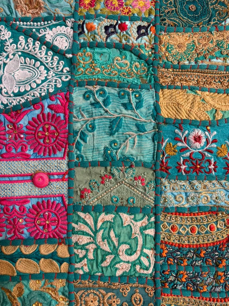 Pode incluir: Um t&ecirc;xtil de patchwork com uma gama vibrante de pain&eacute;is de tecido em tons de azul-petr&oacute;leo, magenta e dourado. A superf&iacute;cie &eacute; densamente decorada com bordados intrincados, fios met&aacute;licos, lantejoulas e padr&otilde;es florais em relevo, unidos por costuras grossas em azul-petr&oacute;leo.