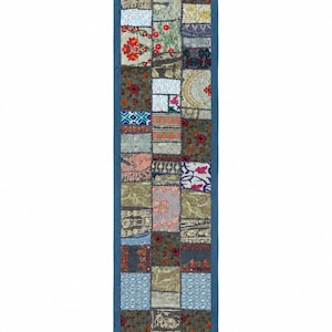 Peut inclure: Un chemin de table rectangulaire composé d'un assemblage de carrés de tissus variés dans des tons de bleu, vert, rouge et doré. Le chemin est bordé d'un cadre en tissu bleu uni et terminé par de petits pompons bleus aux quatre coins.