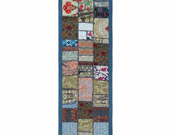 Pode incluir: Um caminho de mesa retangular com um design de retalhos de vários quadrados de tecido em tons de azul, verde, vermelho e dourado. O caminho é contornado por uma moldura de tecido azul sólido e finalizado com pequenas borlas azuis nos cantos.