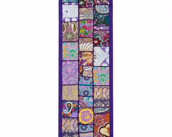 Pode incluir: Um caminho de mesa retangular com um design de patchwork de vários quadrados de tecido em tons de roxo, rosa, amarelo e verde. O caminho é contornado por uma moldura de tecido roxo sólido e finalizado com pequenas borlas roxas em cada um dos quatro cantos.