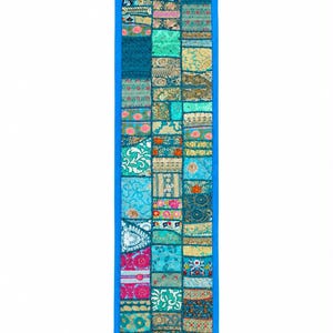 May include: A long, rectangular table runner featuring a patchwork design of various blue, teal, and gold fabric squares with intricate floral and geometric patterns. The runner is bordered by a solid blue fabric and finished with blue tassels at each corner.