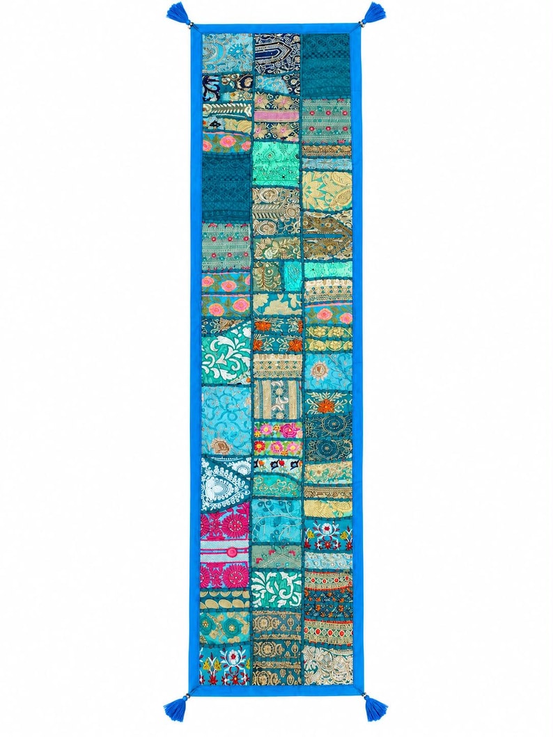 Pode incluir: Um caminho de mesa longo e retangular com um design de patchwork de v&aacute;rios quadrados de tecido azul, turquesa e dourado com padr&otilde;es florais e geom&eacute;tricos intrincados. O caminho &eacute; contornado por tecido azul s&oacute;lido e borlas azuis nos cantos.