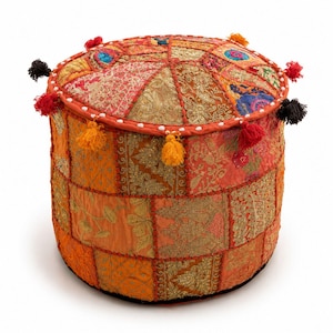Pode incluir: Um pufe redondo com um design de retalhos de painéis de tecido laranja, dourado e vermelho com bordados metálicos intrincados. As bordas são decoradas com pequenas borlas coloridas em vermelho, amarelo e preto, criando uma superfície texturizada.