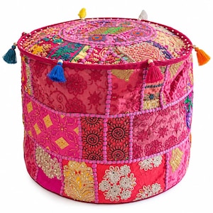 May include: A round, cylindrical ottoman featuring a patchwork design of vibrant pink, magenta, and gold fabrics. The surface is adorned with intricate embroidery, sequins, and floral patterns, finished with colorful tassels along the top edge.