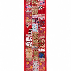 May include: A long, rectangular table runner featuring a vibrant patchwork design of various embroidered fabrics in shades of red, gold, blue, and white. The runner is bordered by a solid red fabric and finished with small red tassels at each corner.