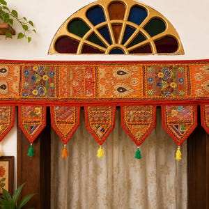 May include: A rectangular orange fabric valance featuring intricate embroidery, mirror work, and patchwork panels. The bottom edge has seven pointed pennant-style drops, each finished with a small, colorful tassel in yellow or green.