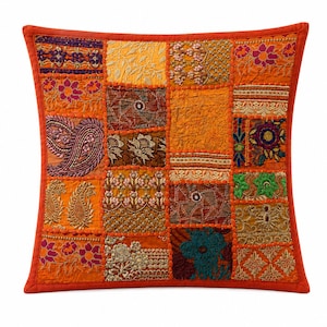 Puede incluir: Un cojín cuadrado con un diseño de patchwork compuesto por varios cuadrados de tela en tonos naranja, dorado y marrón. Cada sección muestra bordados intrincados, patrones florales, motivos de cachemira y detalles en hilo metálico, todo bordeado por un marco naranja sólido.