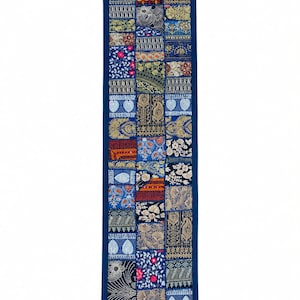 Puede incluir: Un camino de mesa rectangular con un diseño de patchwork de varios cuadrados de tela bordados en azul, dorado y blanco. El camino está bordeado con tela azul oscuro sólida y rematado con pequeñas borlas azul oscuro en las cuatro esquinas.