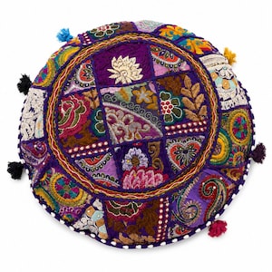 Könnte beinhalten: Ein rundes Bodenkissen mit einem Patchwork-Design aus violetten, magentafarbenen, grünen und gelben Stoffbahnen. Die Oberfläche ist mit aufwendigen Blumenstickereien, Pailletten und kleinen blauen, schwarzen, gelben und rosa Quasten verziert.