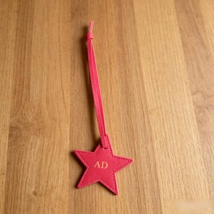 May include: A bright pink star-shaped luggage tag featuring a long, thin matching strap. The center of the star is embossed with the gold-colored capital letters AD. The tag is constructed with visible stitching along the edges of the star shape.