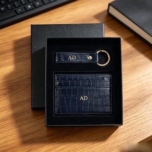 May include: A navy blue crocodile-embossed leather card holder and a matching keychain set presented in a black gift box. Both items feature the gold-embossed initials AD. The keychain includes a gold-toned metal ring and a small gold stud detail.