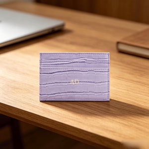 May include: A light purple card holder with a textured crocodile skin pattern. The rectangular accessory features horizontal slots for cards and the gold-colored embossed letters AD centered on the front surface with visible stitching.